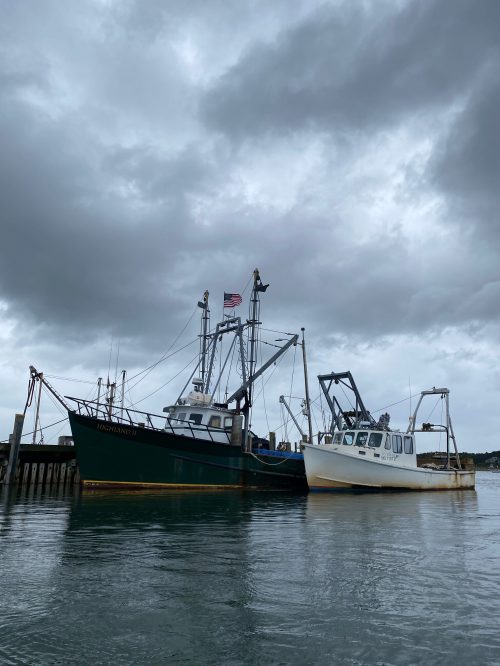 Two fishing boats docked at a pier under a cloudy sky, with one displaying an American flag.