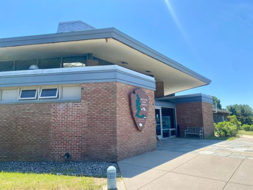 A modern brick building with large windows and a sign, surrounded by greenery under a clear blue sky.