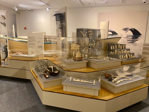 Display of various artifacts in a museum, including carved items and a traditional boat in the background.
