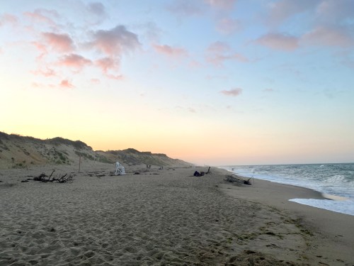 A serene beach at sunset, with gentle waves, sandy shore, and distant dunes under a colorful sky.
