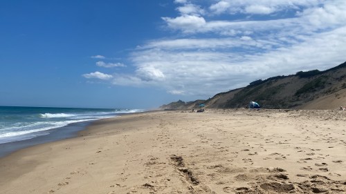 A sandy beach with gentle waves, dunes in the background, and a clear blue sky with scattered clouds.