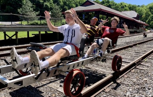 Three young men ride a rail bike on a sunny day, smiling and waving, with a scenic background of trees and a building.