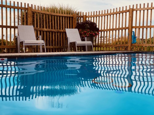 A serene pool scene with two lounge chairs beside the water, surrounded by a wooden fence and lush greenery.
