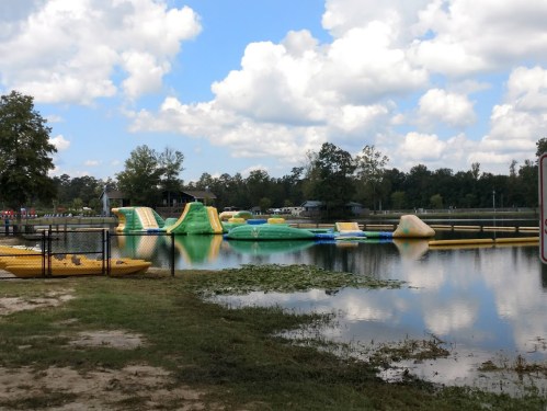 A water park with inflatable structures on a lake, surrounded by trees and a cloudy sky. Kayaks are visible in the foreground.