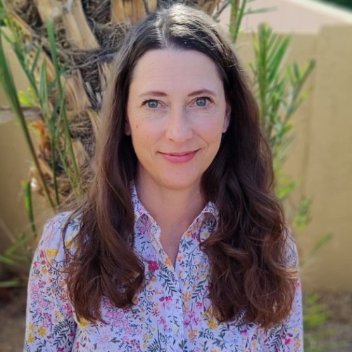 A woman with long brown hair smiles, wearing a floral shirt, with greenery in the background.