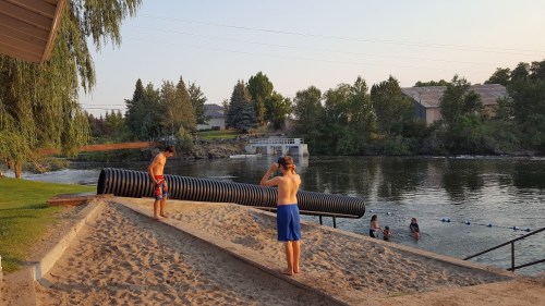 Two boys in swim trunks stand on a sandy area by a river, with others swimming in the background and trees lining the shore.