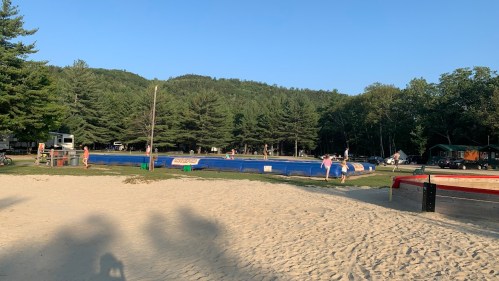 A sandy area with a blue inflatable pool, surrounded by trees and campers in the background under a clear blue sky.