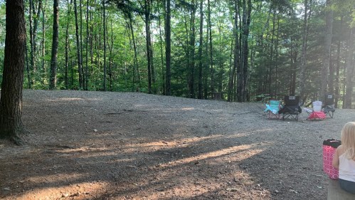 A serene forest scene with two camping chairs set up on a dirt area, surrounded by tall trees and dappled sunlight.