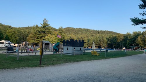 A playground with a castle structure, surrounded by trees and mountains, with a gravel path leading through the area.