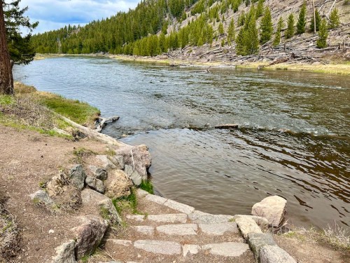 A serene riverbank with stone steps leading down to a calm river, surrounded by lush green trees and a rocky landscape.
