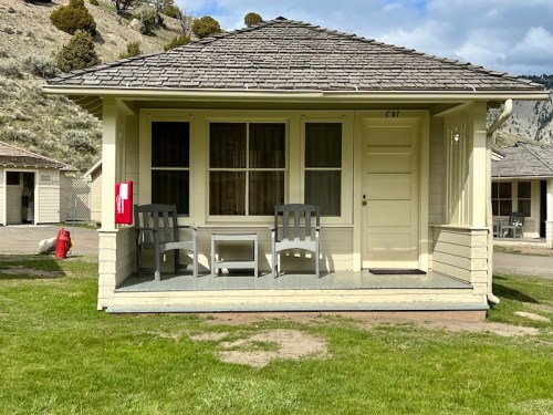 A small cabin with a porch, two chairs, and a fire extinguisher nearby, set against a grassy landscape and hills.