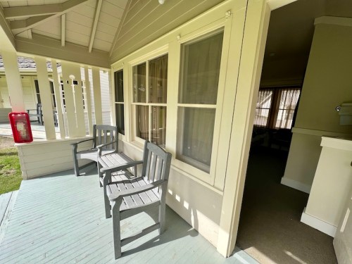 A porch with two wooden chairs and a fire extinguisher, leading to a room with large windows.