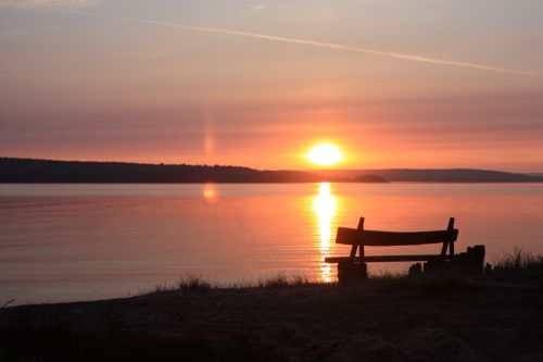 A serene sunset over calm waters, with a silhouette of a bench in the foreground.