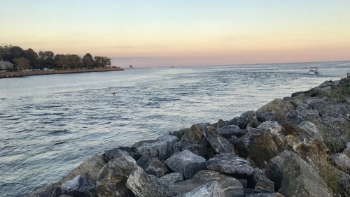 A serene waterfront scene with rocks in the foreground and a calm river under a pastel sunset sky.