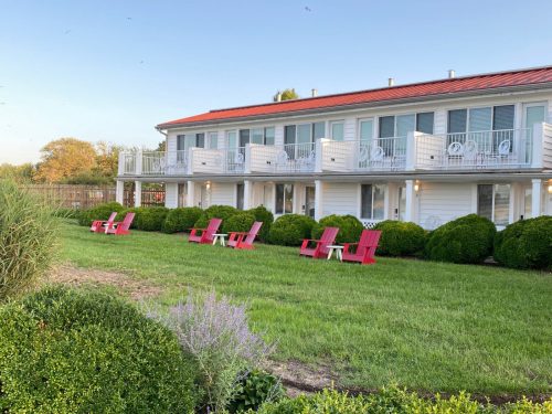 A two-story building with balconies, surrounded by green grass and neatly trimmed bushes, featuring pink lawn chairs.