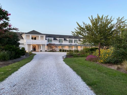 A spacious white house with a gravel driveway, surrounded by lush gardens and trees under a clear blue sky.
