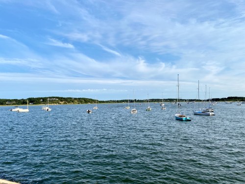 A serene view of a calm bay with several sailboats anchored under a blue sky with wispy clouds.