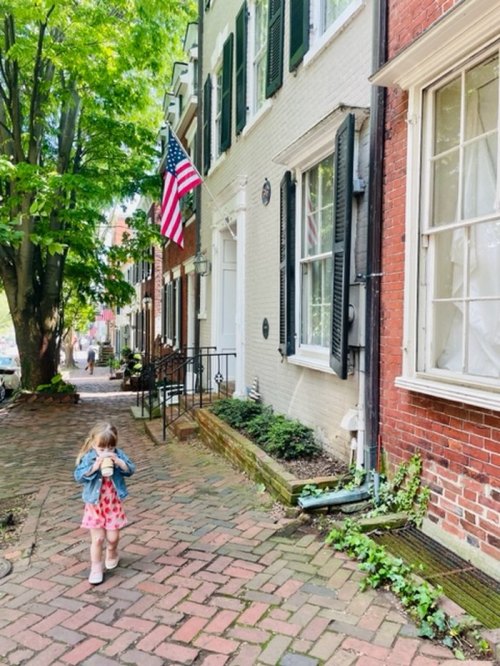 A child in a pink dress walks down a brick path lined with trees and historic houses, with an American flag visible.
