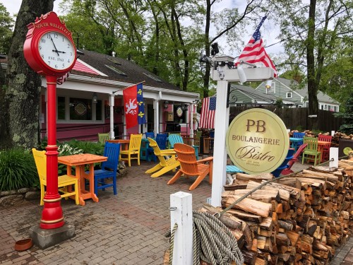 Colorful outdoor seating at a bistro with a clock, flags, and stacked firewood in a charming setting.