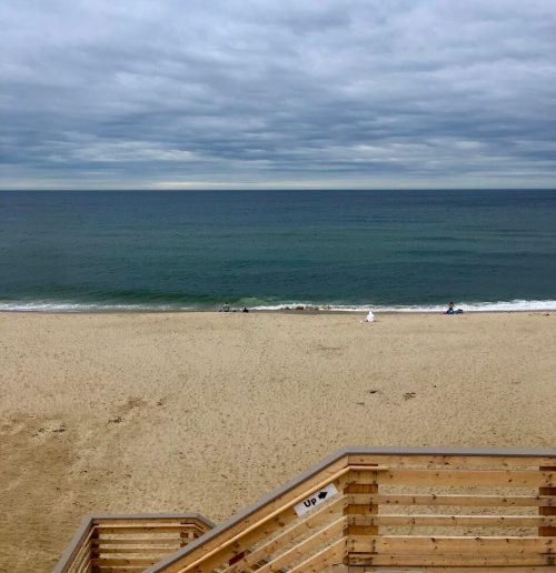 A sandy beach with gentle waves under a cloudy sky, viewed from a wooden deck.