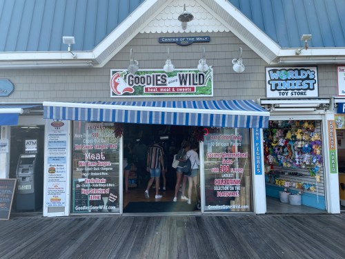 A storefront with a striped awning, advertising meat and sweets, next to a toy store on a boardwalk.
