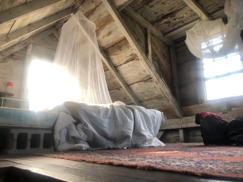 A rustic attic room with a bed draped in white fabric, sunlight streaming through a window, and scattered clothing.