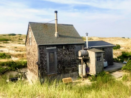 A rustic wooden house with a chimney, surrounded by sandy dunes and grass under a clear blue sky.