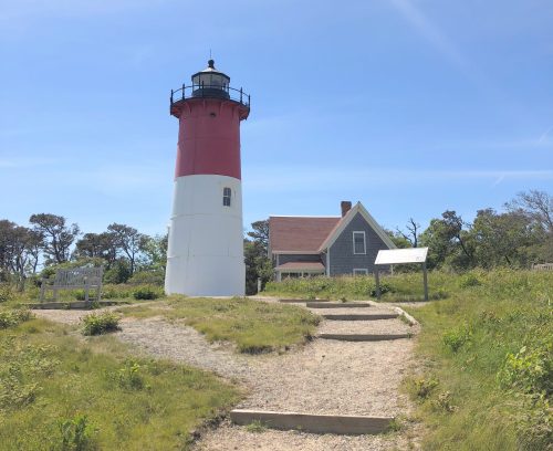 A red and white lighthouse stands beside a gray house, surrounded by green grass and a clear blue sky.