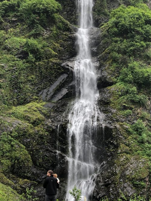 A person holds a child while standing in front of a tall waterfall surrounded by lush greenery.