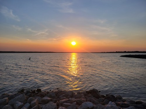 A serene sunset over a calm lake, with golden reflections on the water and rocky shoreline in the foreground.