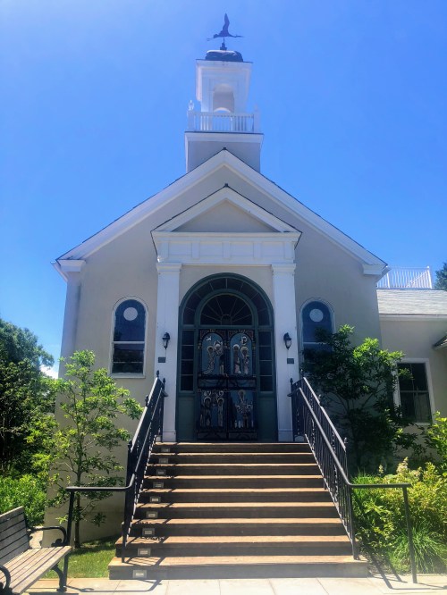 A charming church with a white facade, large front doors, and a steeple, surrounded by greenery under a clear blue sky.