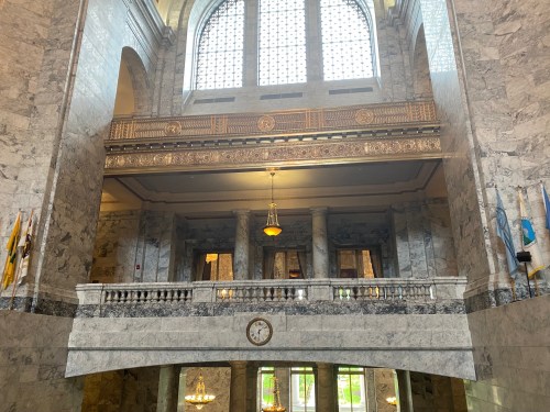 Interior view of a grand marble hall with large windows, ornate railings, and a clock on the wall.