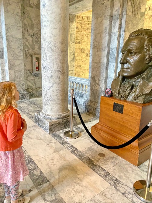 A young girl in a pink dress and orange jacket gazes at a large bronze bust in a marble hall.