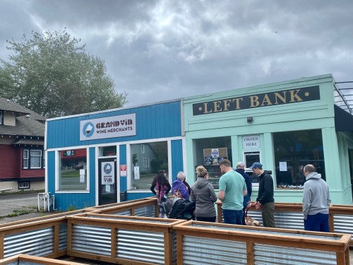 A group of people gathers outside two storefronts, one labeled "Grand Vin Wine Merchants" and the other "Left Bank."