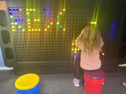 A child sits on a stool, playing with colorful lights on a black pegboard, creating patterns and shapes.