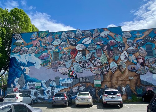 Colorful mural on a building featuring various painted leaves and ocean scenes, with parked cars in the foreground.