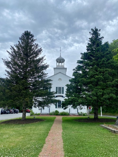 A path leads to a white building with a steeple, flanked by tall evergreen trees under a cloudy sky.