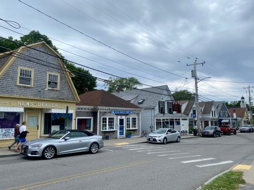 A quaint street lined with shops and cars, under a cloudy sky, showcasing a small-town atmosphere.