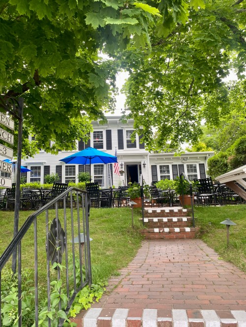 A charming house with a brick pathway, surrounded by greenery and outdoor seating under blue umbrellas.