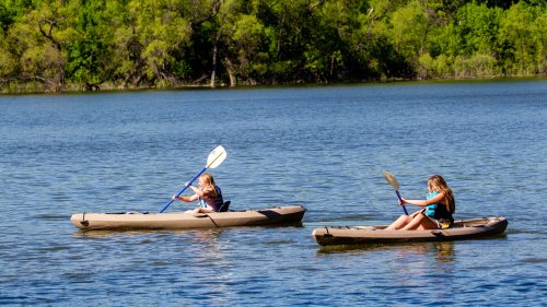 Two girls kayaking on a calm lake, surrounded by lush green trees.