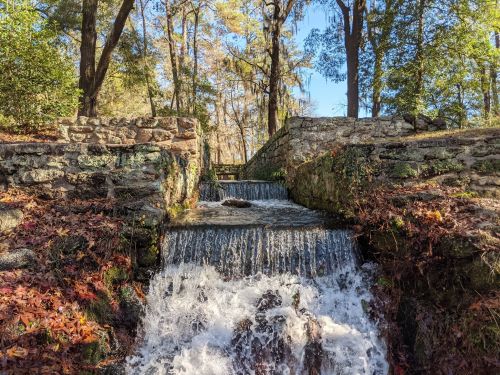 A small waterfall cascades over stone steps, surrounded by trees and autumn leaves in a serene outdoor setting.