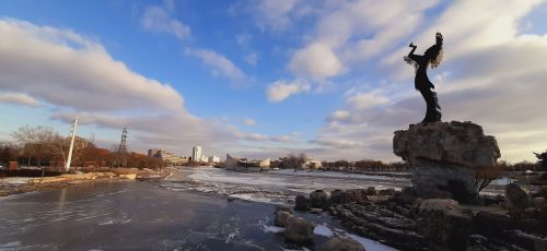A silhouette of a statue on a rock by a frozen river, with a cloudy blue sky and city skyline in the background.