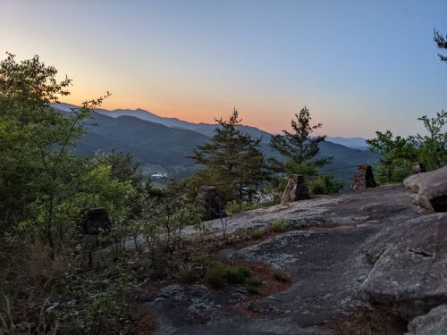 A serene mountain landscape at sunset, with rocky outcrops and trees framing the view of distant hills.