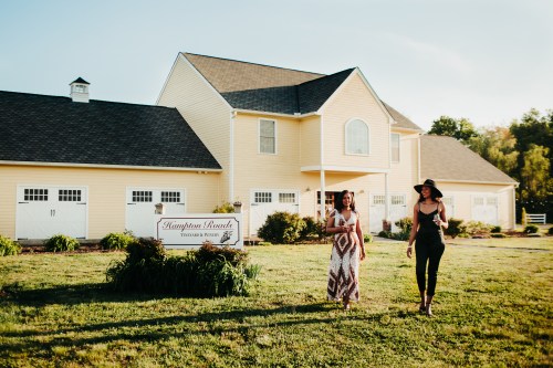 Two women walk towards a yellow house with a sign that reads "Hamptons Ranch," surrounded by greenery.