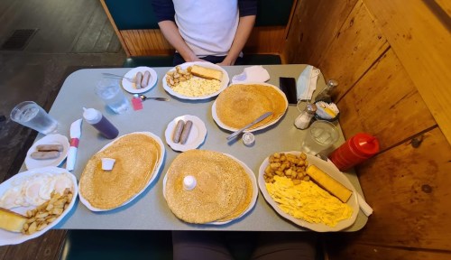 A table set with multiple plates of pancakes, scrambled eggs, sausage links, and hash browns, with drinks nearby.