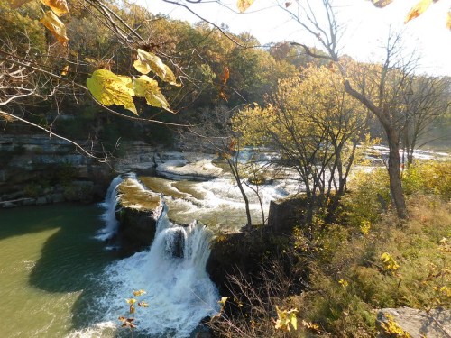 A serene waterfall cascades into a river, surrounded by autumn foliage and rocky cliffs under a clear blue sky.