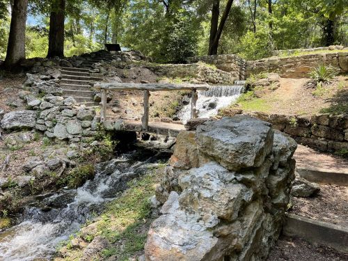 A serene landscape featuring a small wooden bridge over a flowing stream, surrounded by lush greenery and stone pathways.