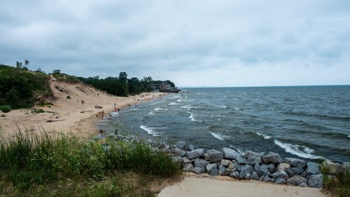 A sandy beach with gentle waves, rocky shoreline, and overcast skies, surrounded by greenery and distant buildings.