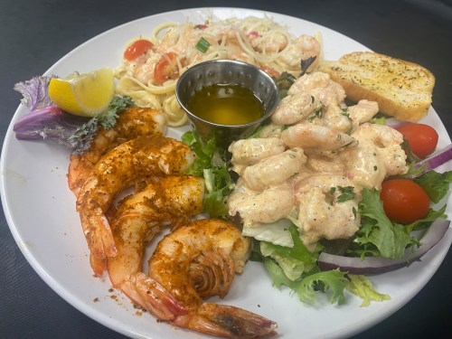 A plate featuring grilled shrimp, creamy shrimp salad, pasta, garlic bread, and fresh salad with tomatoes and lemon.