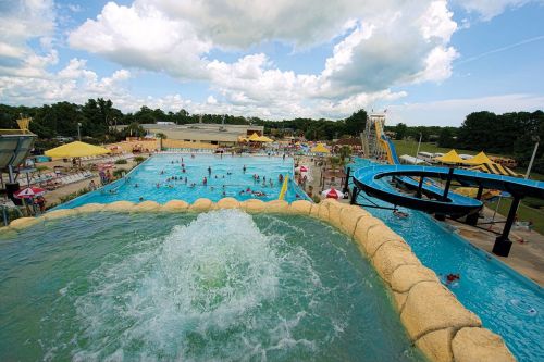A vibrant water park scene with pools, slides, and people enjoying the sunny day under a partly cloudy sky.